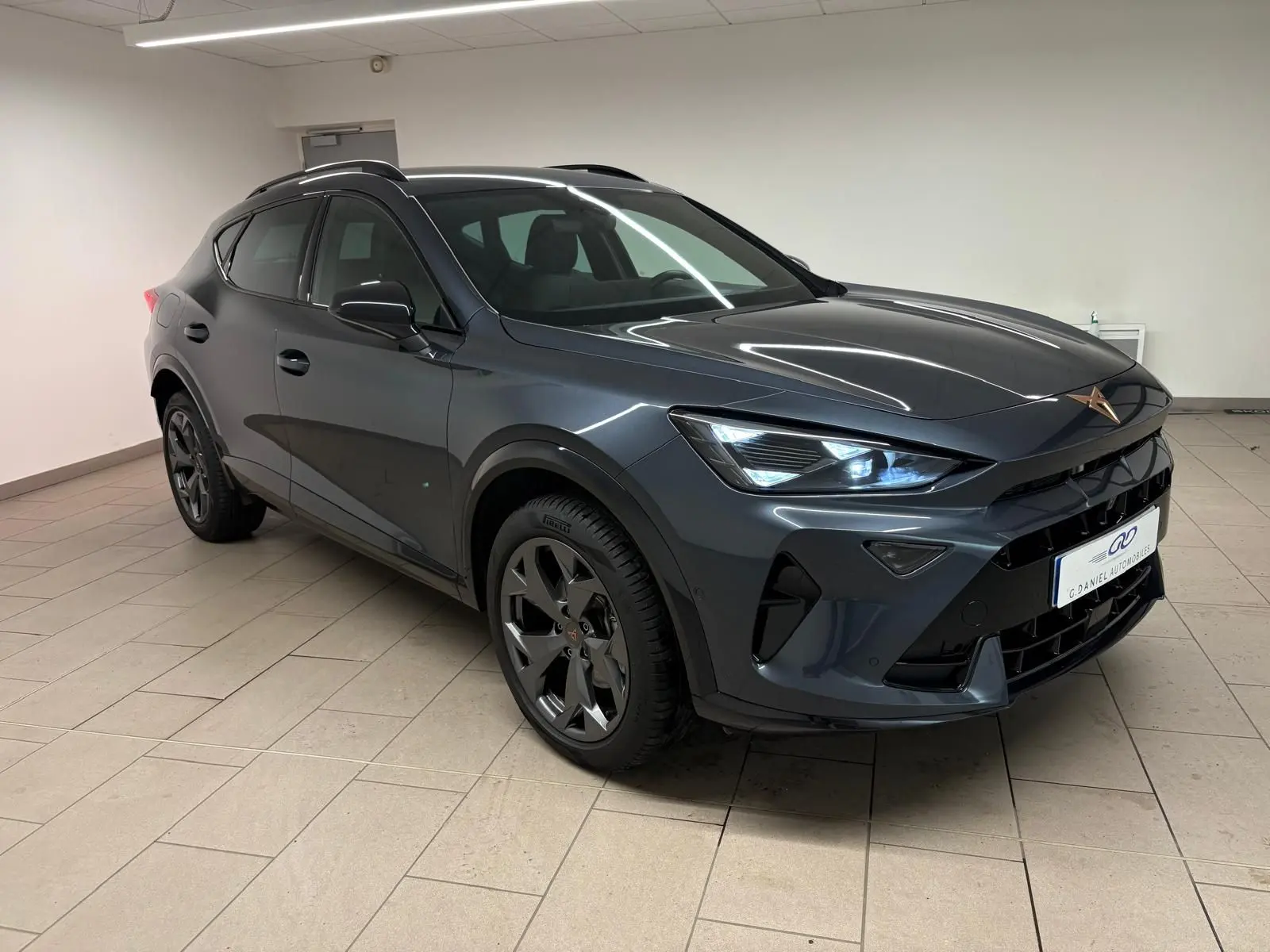 Vue 3/4 avant droit d'un CUPRA Formentor gris magnétique avec phares allumés et jantes noires en intérieur showroom.
