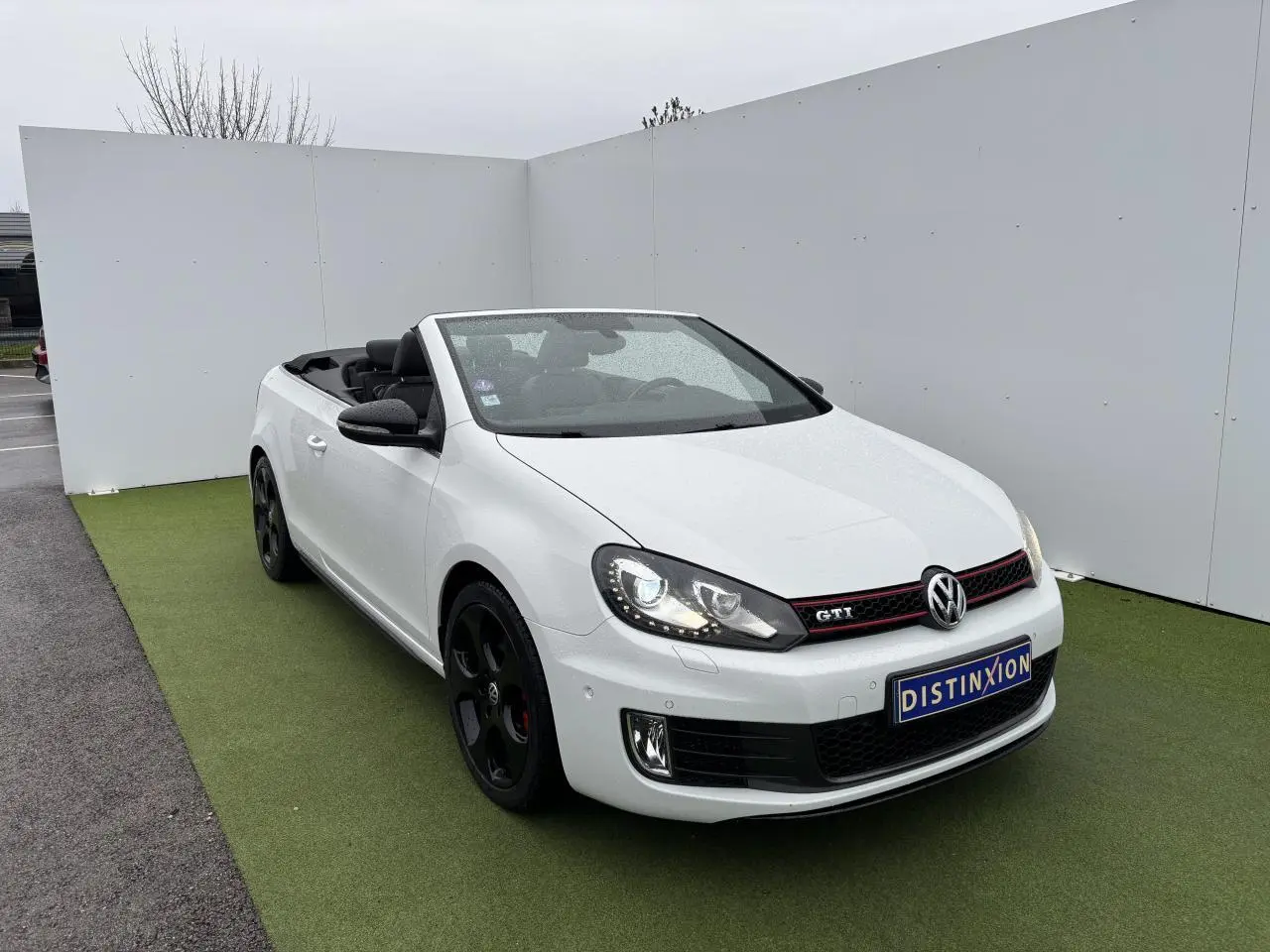 Volkswagen Golf VI Cabriolet blanc vue 3/4 avant avec jantes noires et toit ouvert sur fond neutre.