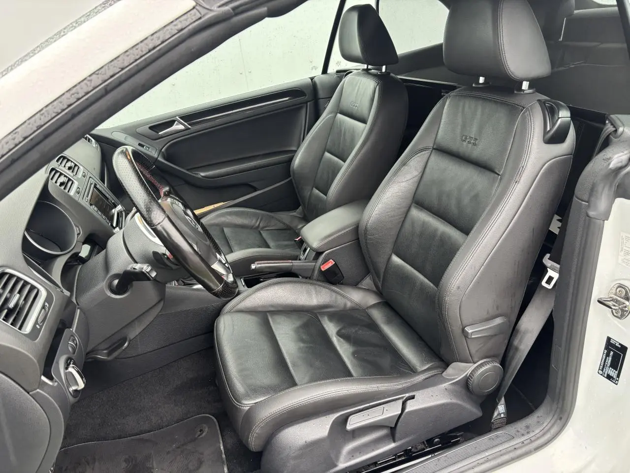 Vue intérieure côté conducteur de la Volkswagen Golf VI Cabriolet blanche, sièges sport en cuir noir avec logo GTI brodé.