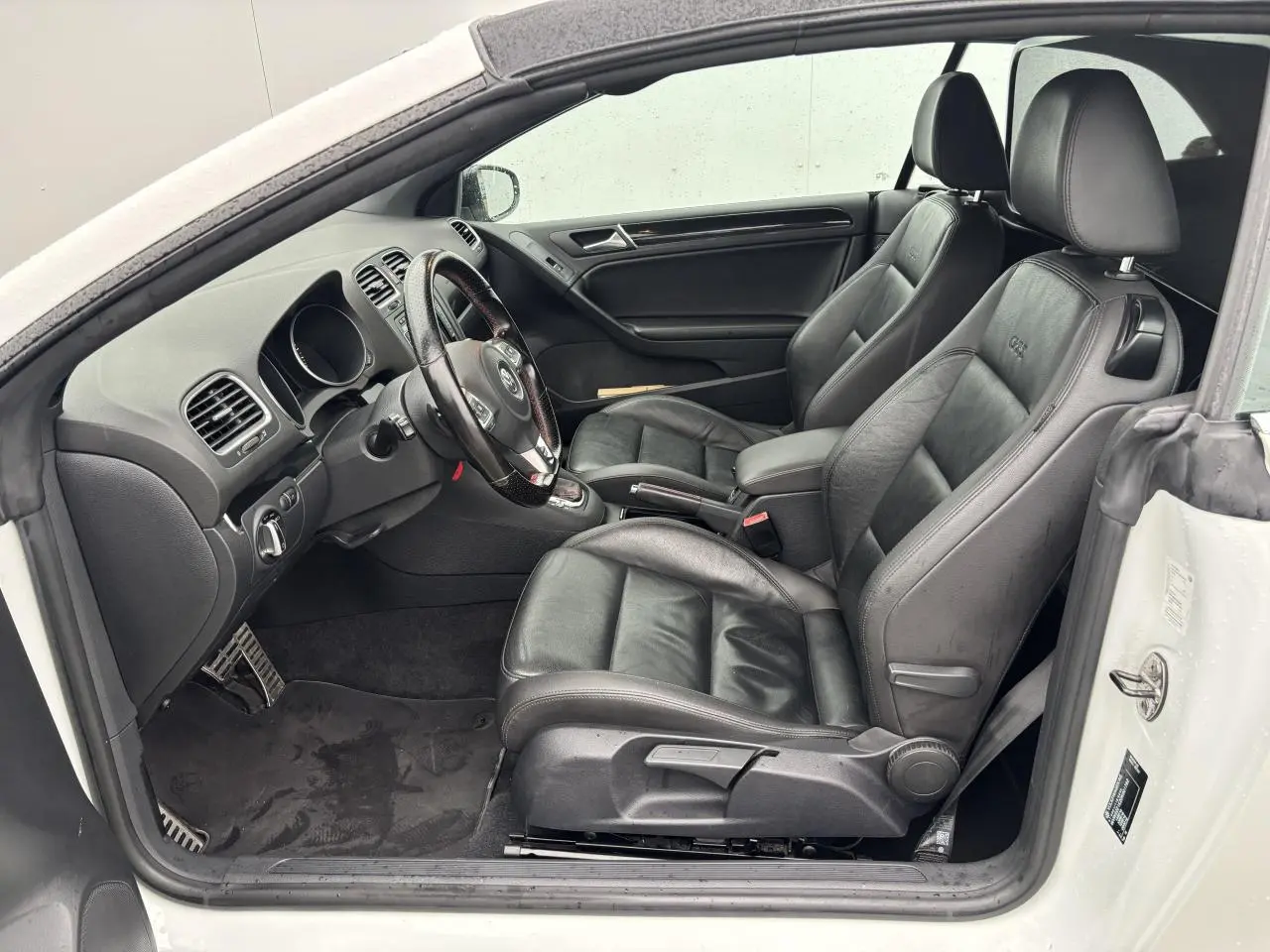 Intérieur côté conducteur d'une Volkswagen Golf VI Cabriolet blanc, sièges sport cuir noirs avec logo GTI brodé.