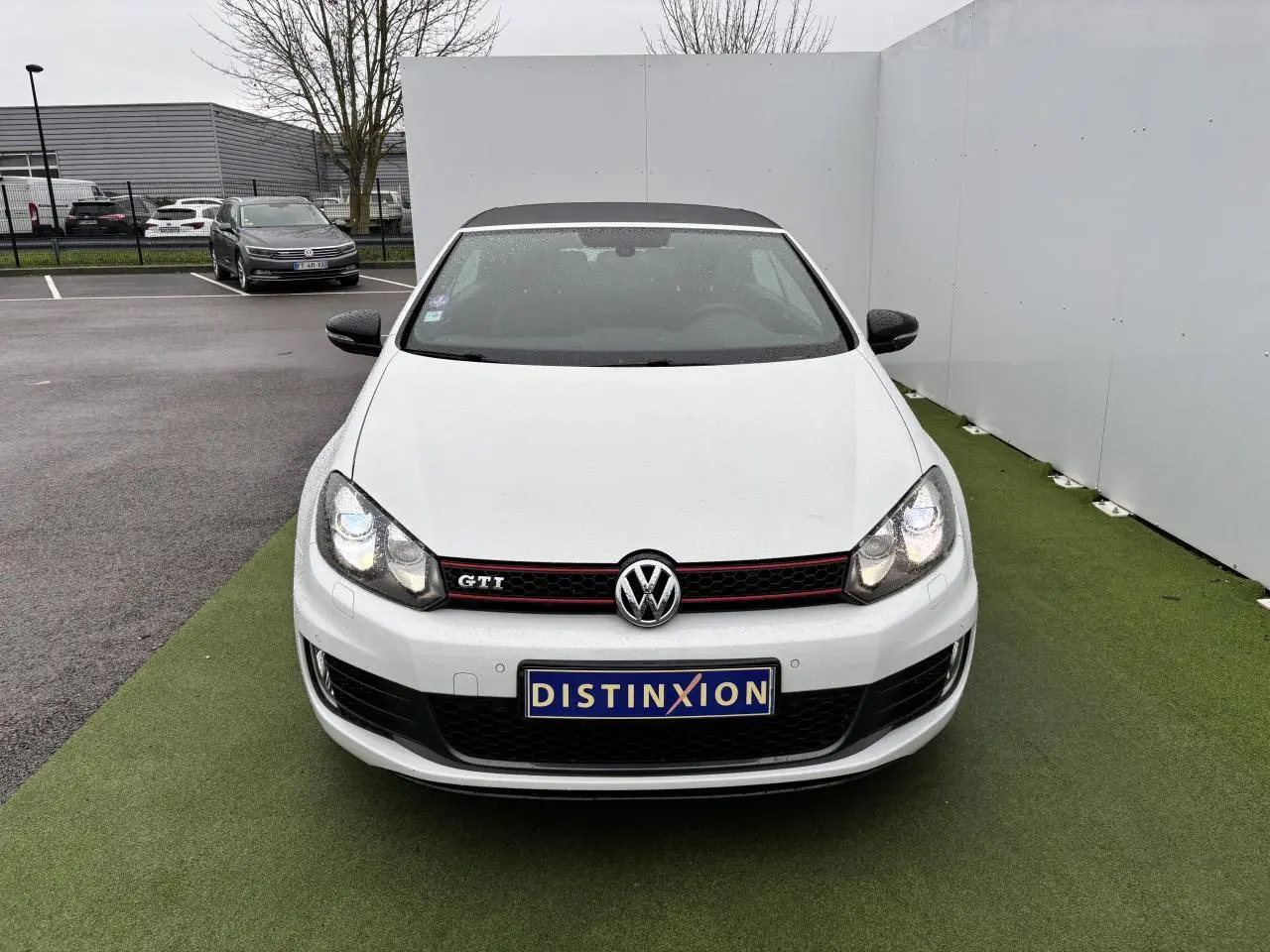 Vue avant d'une Volkswagen Golf VI Cabriolet GTI blanche avec calandre nid d'abeille et logo GTI rouge.