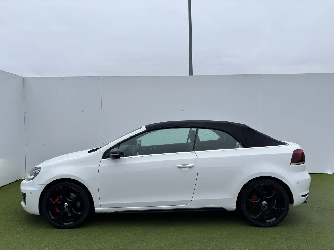Profil côté gauche d'un Volkswagen Golf VI Cabriolet blanc avec toit souple noir et jantes alliage noires.