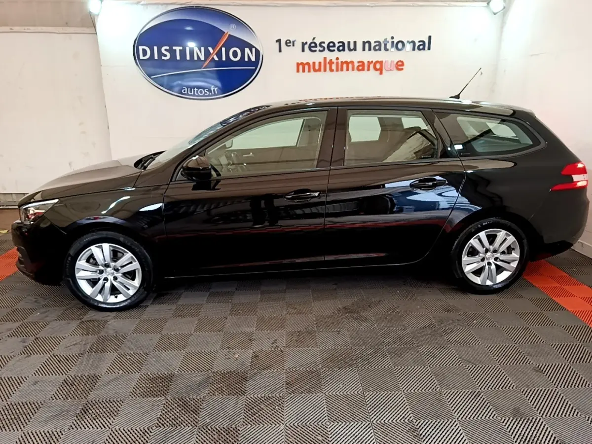 Profil droit d'une Peugeot 308 noire break de 2020, avec jantes alliage et feux arrière allumés en intérieur showroom.