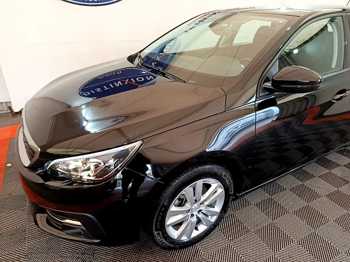 PEUGEOT 308 noir vue 3/4 avant droit, avec phares allumés et jantes alliage argentées visibles.