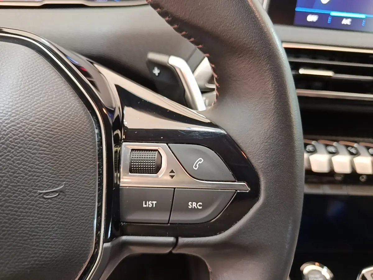 Gros plan sur les commandes gauche du volant cuir noir du Peugeot 3008 2022, avec tableau de bord et écran tactile en arrière-plan.