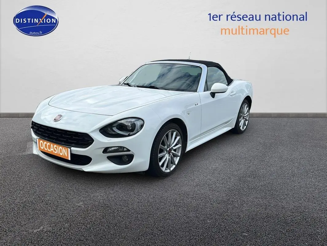 Fiat 124 Spider blanc vue 3/4 avant avec capote noire et jantes alliage 17 pouces sur sol gris.