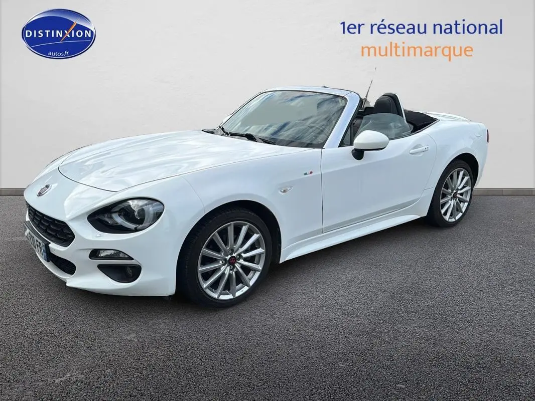 Fiat 124 Spider blanc vue 3/4 avant droit, capote ouverte, jantes alliage 17 pouces et phares antibrouillard visibles.