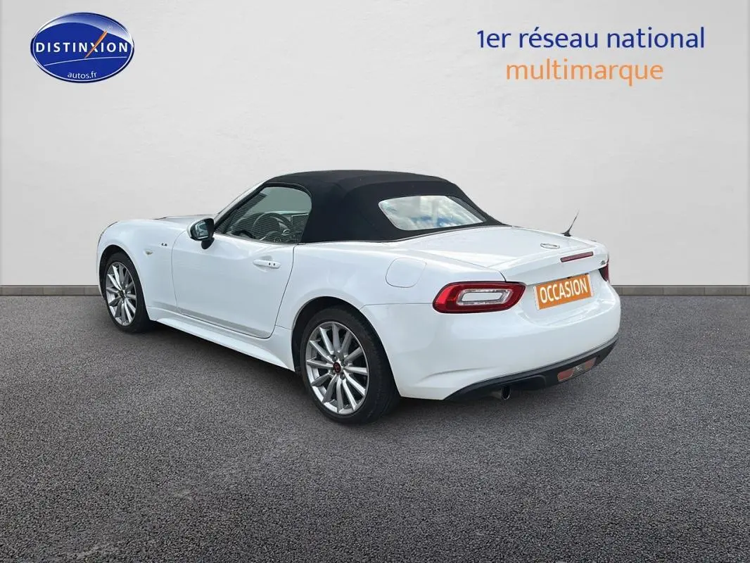 Fiat 124 Spider blanc vue 3/4 arrière côté gauche, capote noire fermée et jantes alliage 17 pouces visibles.