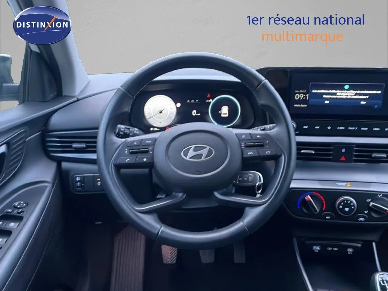 Vue intérieure centrée sur le volant cuir et tableau de bord numérique de la Hyundai i20 blanche, avec commandes au volant visibles.