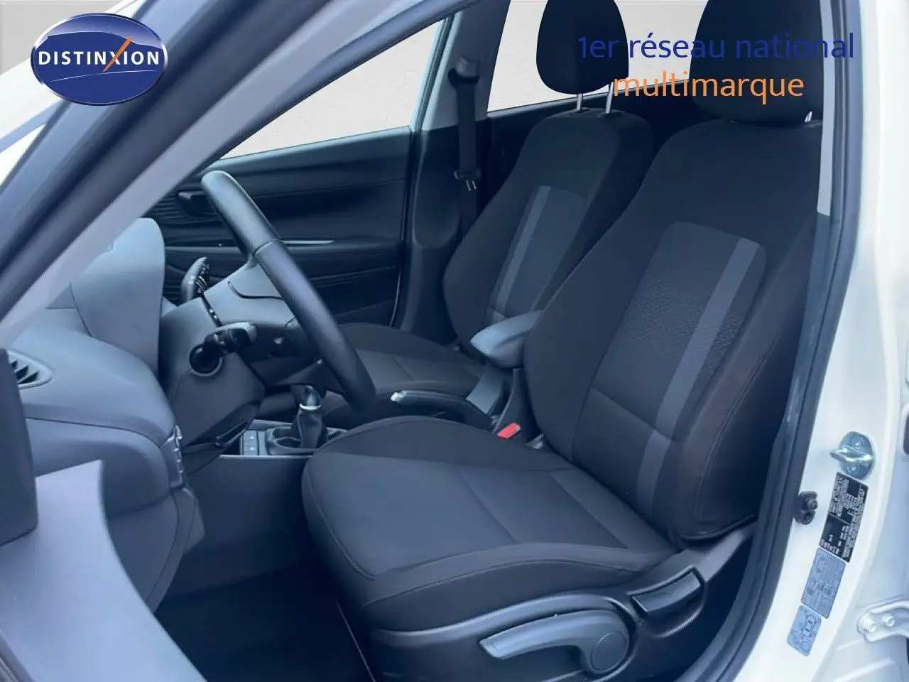 Vue intérieure côté conducteur d'une Hyundai i20 blanche, montrant les sièges noirs en tissu et la boîte manuelle.