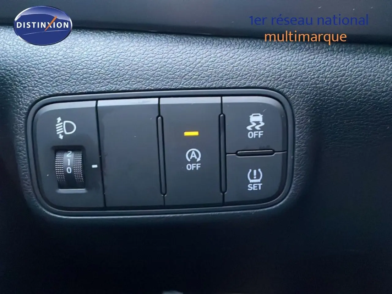 Gros plan sur les boutons de contrôle noirs du tableau de bord d’une Hyundai i20 blanche, avec réglage de phares et aides à la conduite.