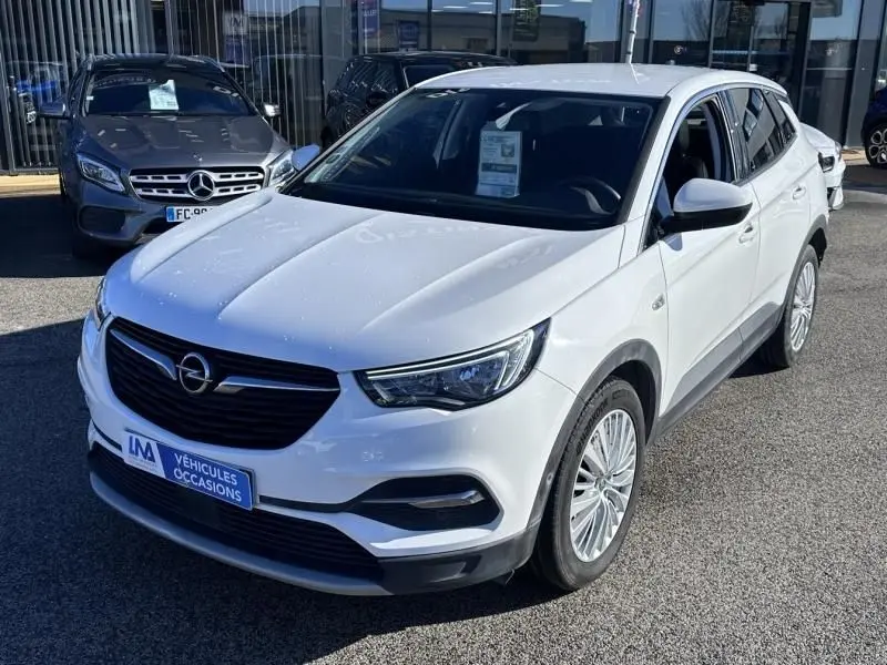 Vue 3/4 avant d’un Opel Grandland X blanc 2018, avec calandre noire et jantes alliage, stationné devant un concessionnaire.
