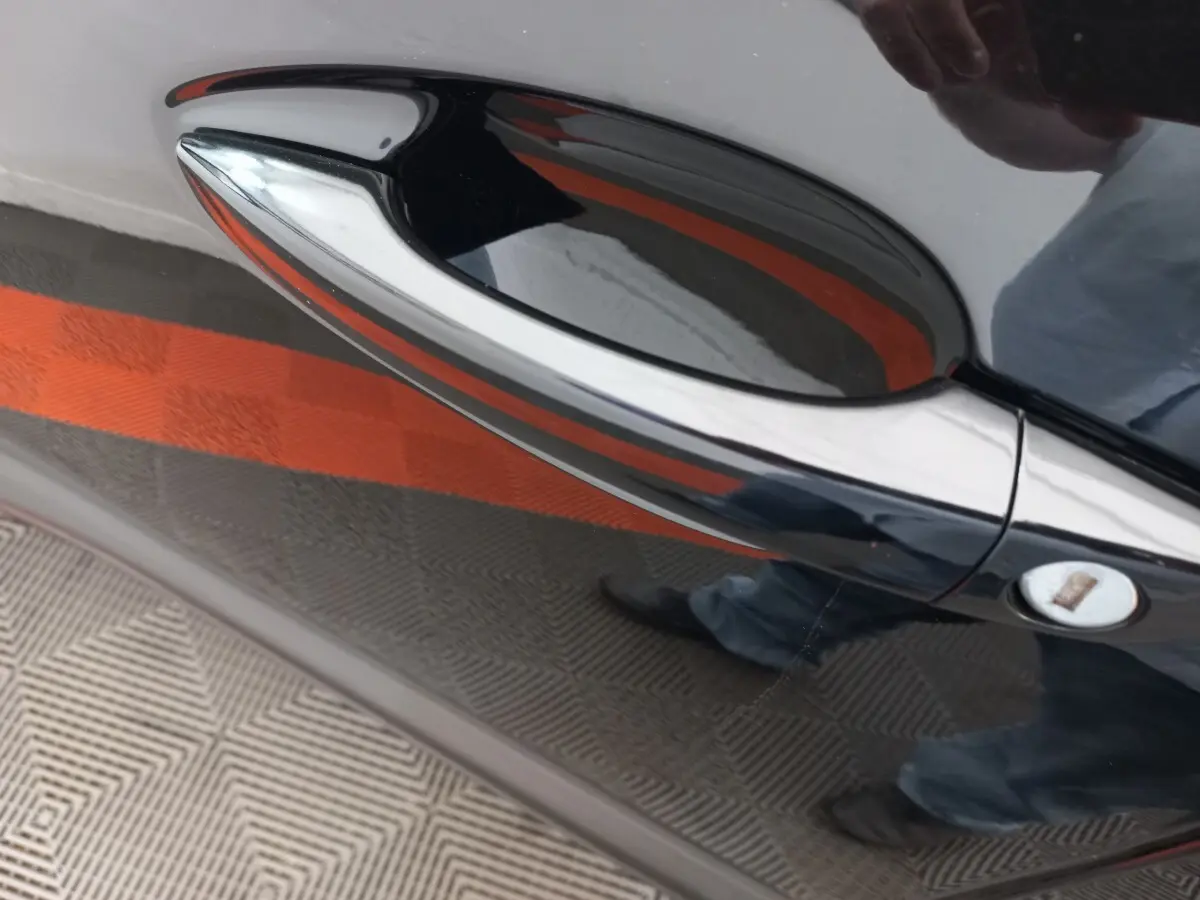 Poignée de porte côté droit noire brillante du Peugeot 3008 PureTech 130, reflet du sol et d'une personne visible.