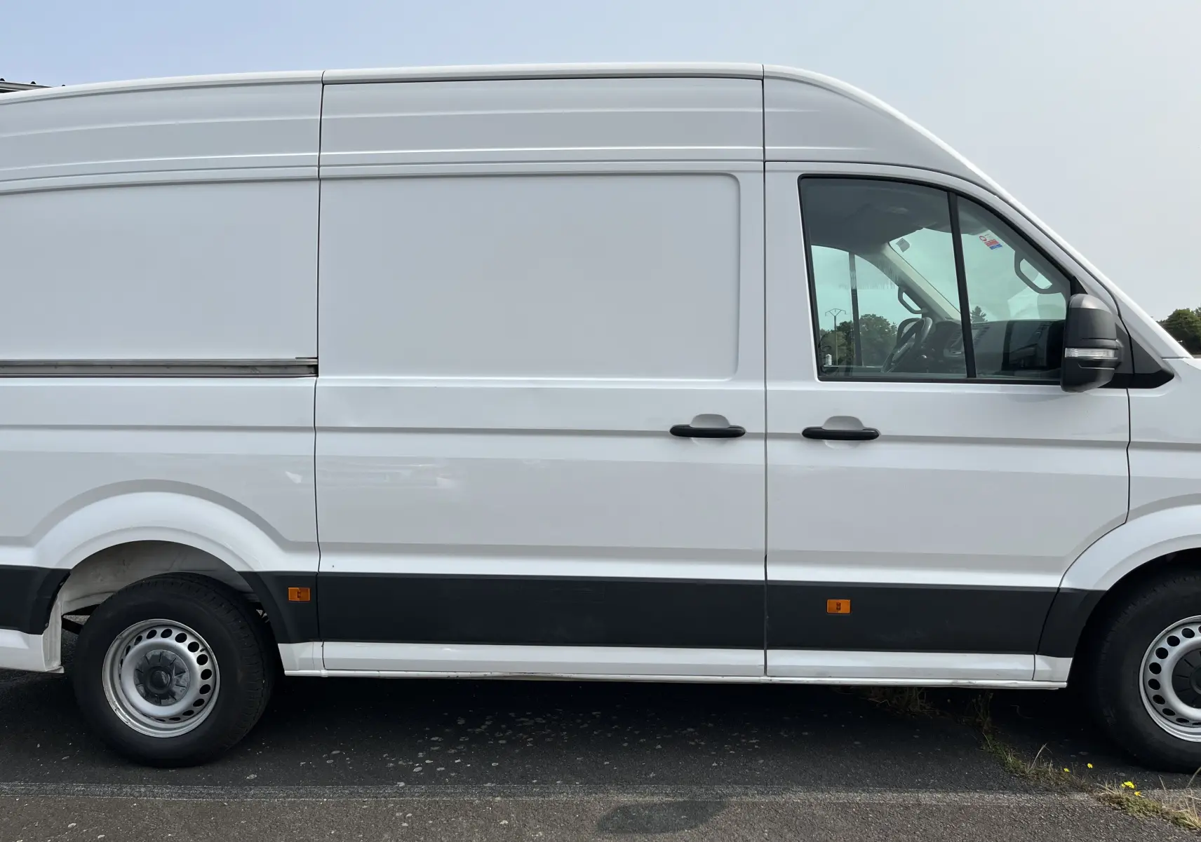 Profil côté gauche d'un Volkswagen Crafter blanc, fourgon utilitaire avec bande noire et feux latéraux orange visibles.