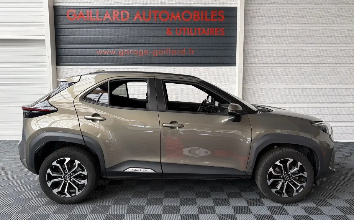 Profil droit d'une Toyota Yaris Cross 2024 gris clair avec jantes alliage noires et logo Yaris Cross sur la porte.