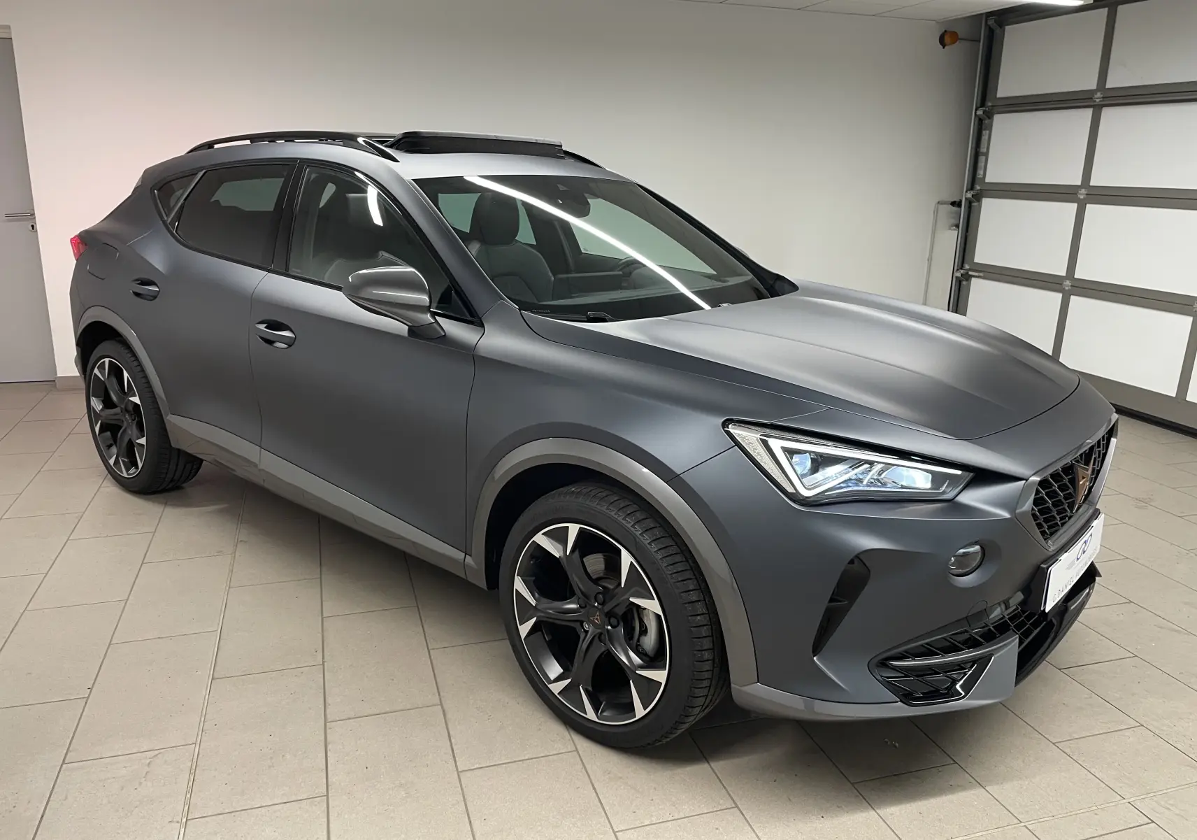 Vue 3/4 avant droite du CUPRA Formentor 1.5 TSI 150 DSG7 en gris magnétique mat avec jantes noires et toit ouvrant.
