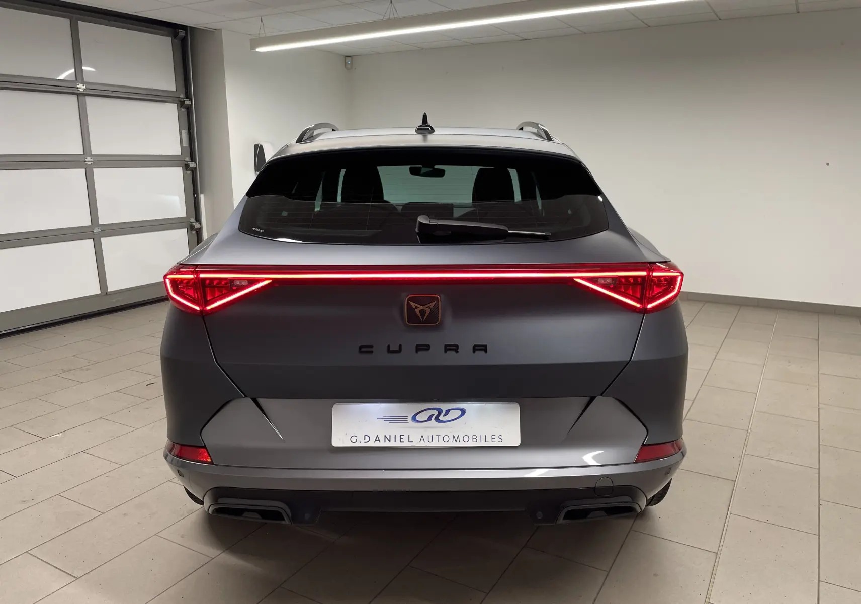 Vue arrière d'un CUPRA Formentor 1.5 TSI 150 DSG7 en gris magnétique mat avec feux LED allumés dans un garage.