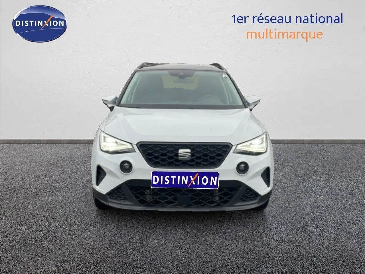 Vue frontale d’un SEAT Arona blanc alpin métal avec toit noir, phares allumés et calandre noire distinctive.
