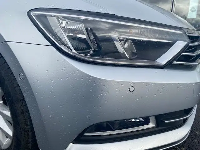Gros plan sur le phare avant gauche argenté d'une Volkswagen Passat 2015 avec gouttes de pluie visibles.