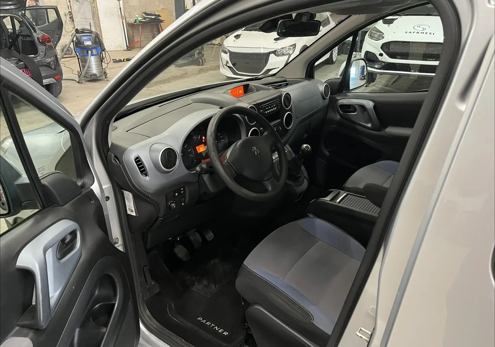 Vue intérieure côté conducteur du Peugeot Partner Tepee gris aluminium, montrant le tableau de bord et les sièges tissu bicolores.