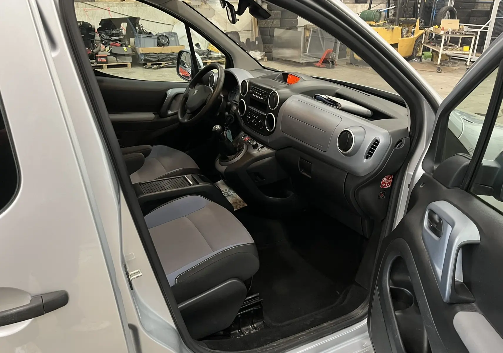 Vue intérieure côté conducteur d'un Peugeot Partner Tepee gris aluminium, montrant le tableau de bord et les sièges en tissu bicolore.