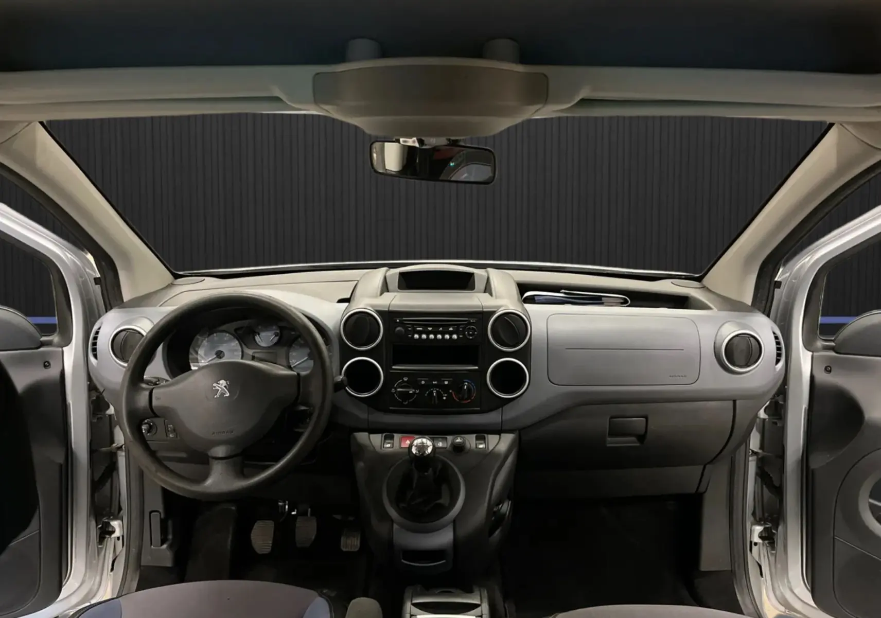 Vue frontale de l’intérieur gris du Peugeot Partner Tepee 2013 avec volant, tableau de bord et portes ouvertes.