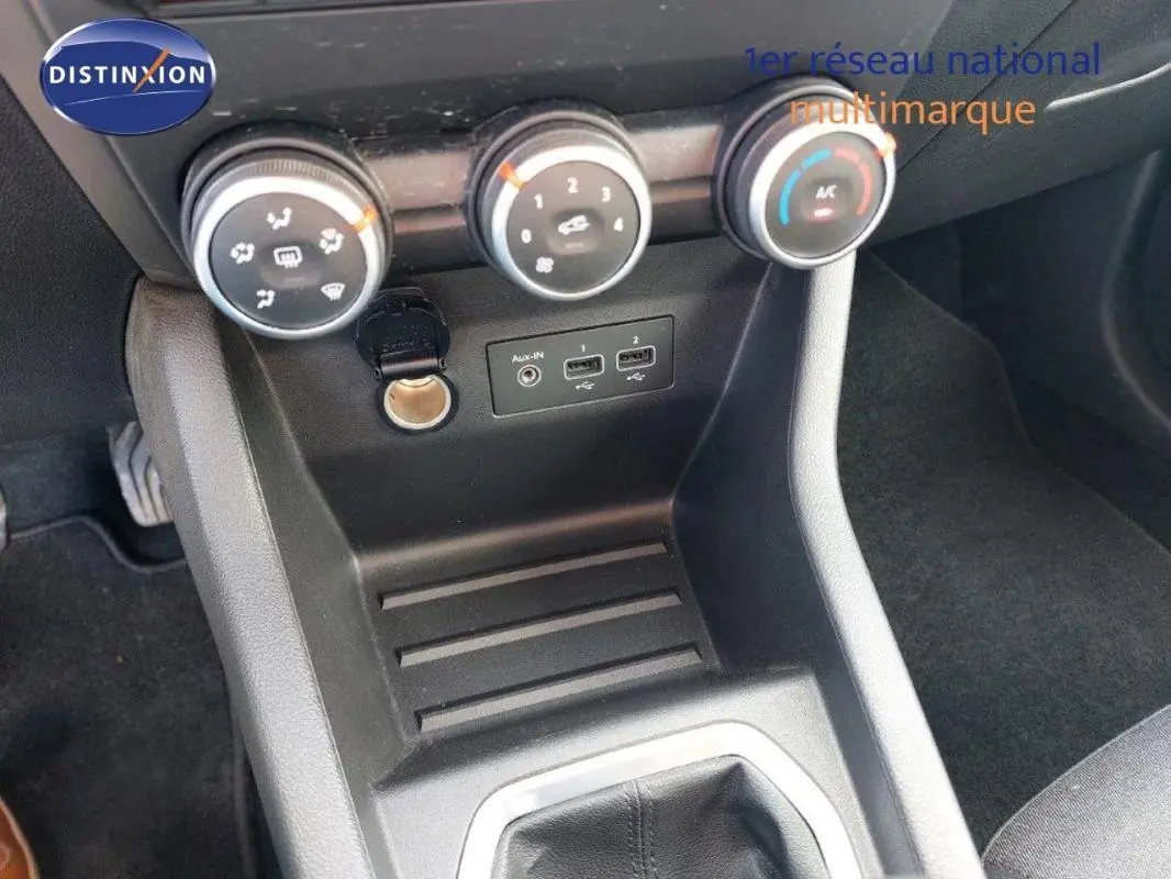 Vue intérieure centrée sur la console centrale de la Renault Clio blanche, avec commandes de climatisation et prise auxiliaire visibles.