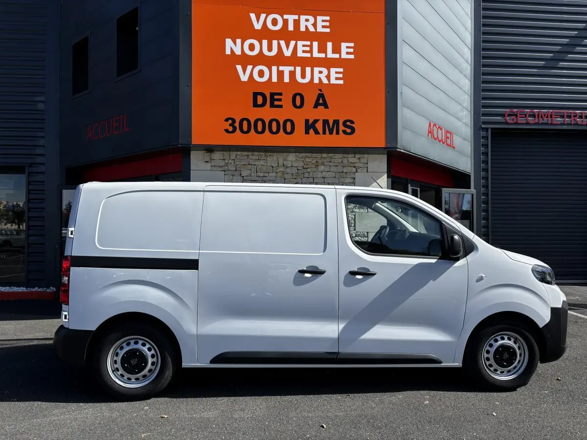 Profil côté droit du Peugeot Expert Fourgon blanc 2025 garé devant un bâtiment avec panneau orange.
