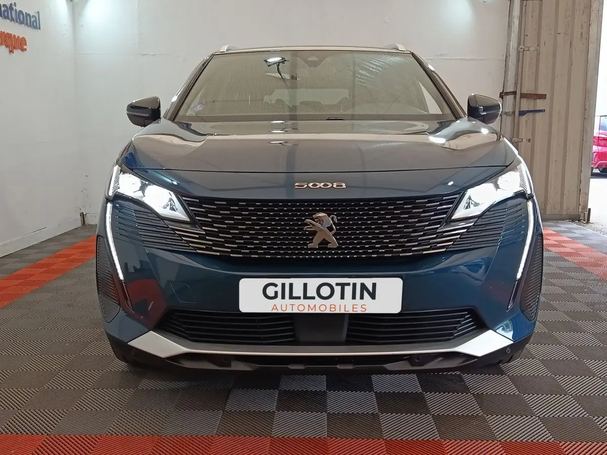 Vue avant d'un Peugeot 5008 bleu 2023 avec calandre noire et feux LED allumés en intérieur showroom.