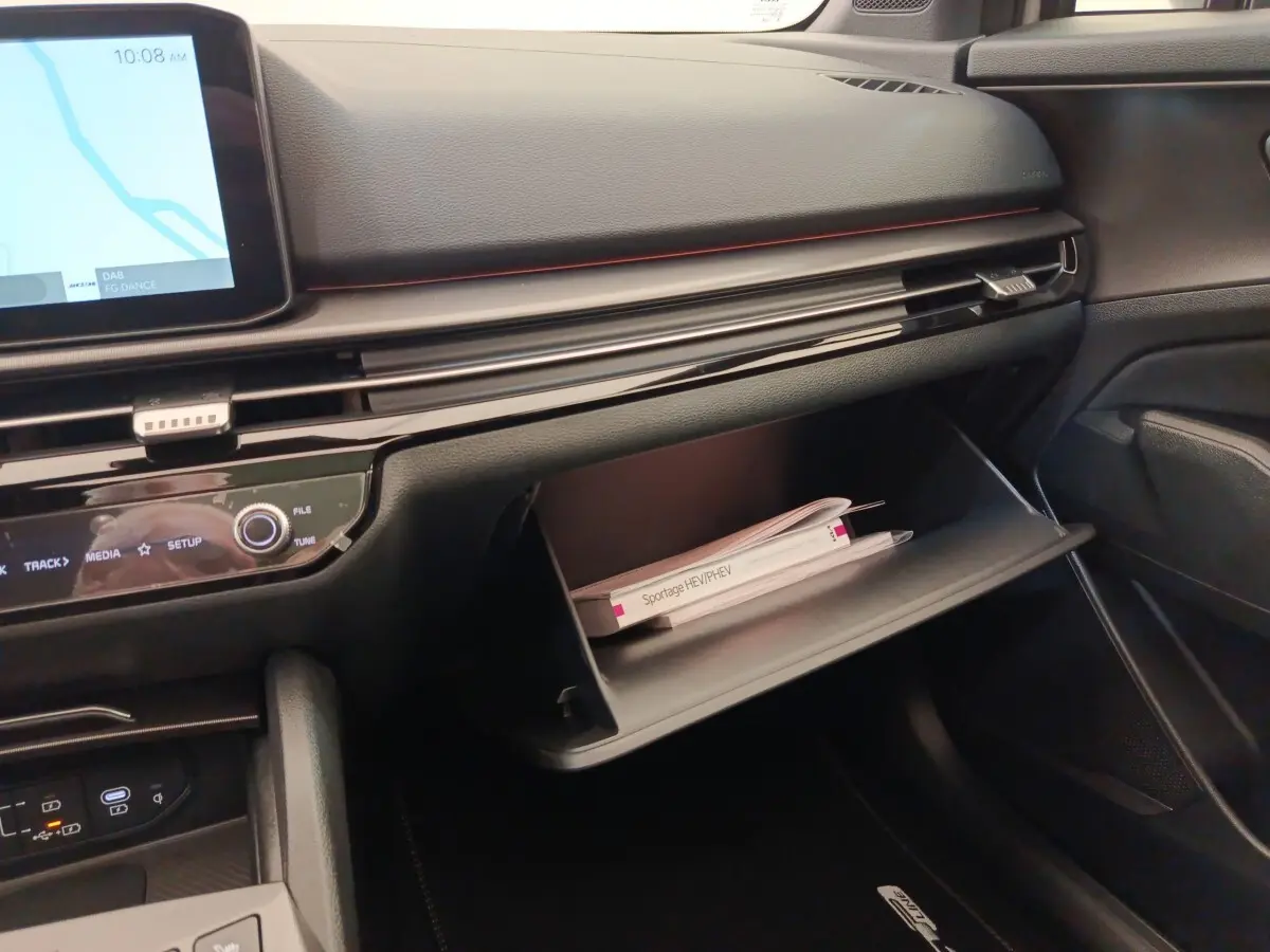 Intérieur KIA Sportage 2026, vue côté passager sur tableau de bord noir avec boîte à gants ouverte et manuel visible.