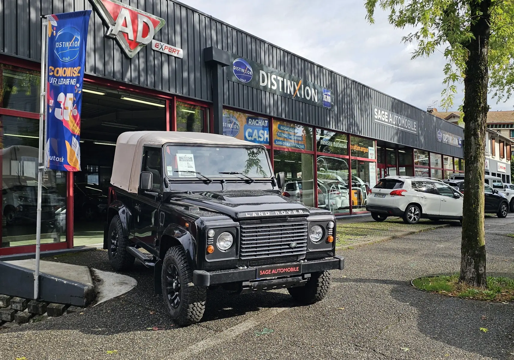 Land Rover 90 Defender noir 3/4 avant avec toit souple beige, stationné devant un garage automobile.