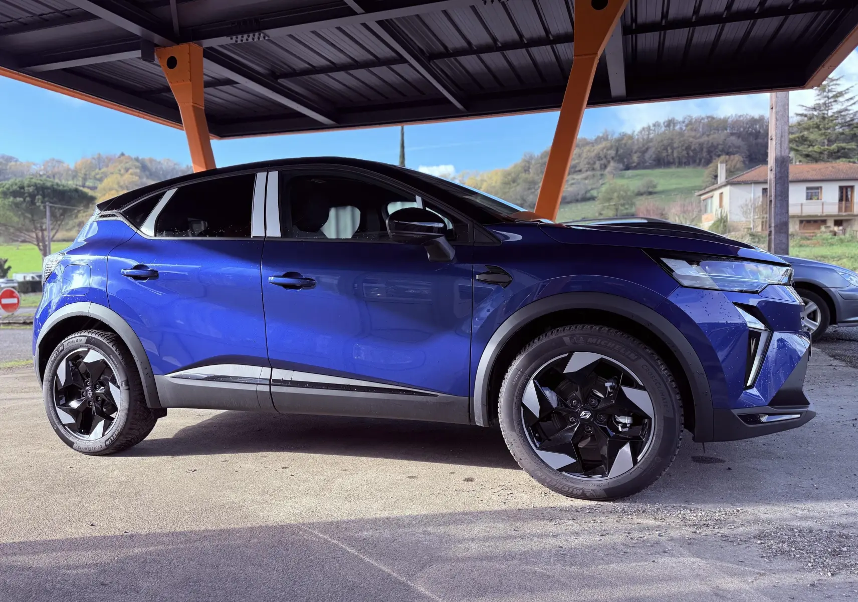 Profil côté gauche du Renault Captur E-Tech hybride 2025 en bleu Iron Metal avec toit noir sous abri.