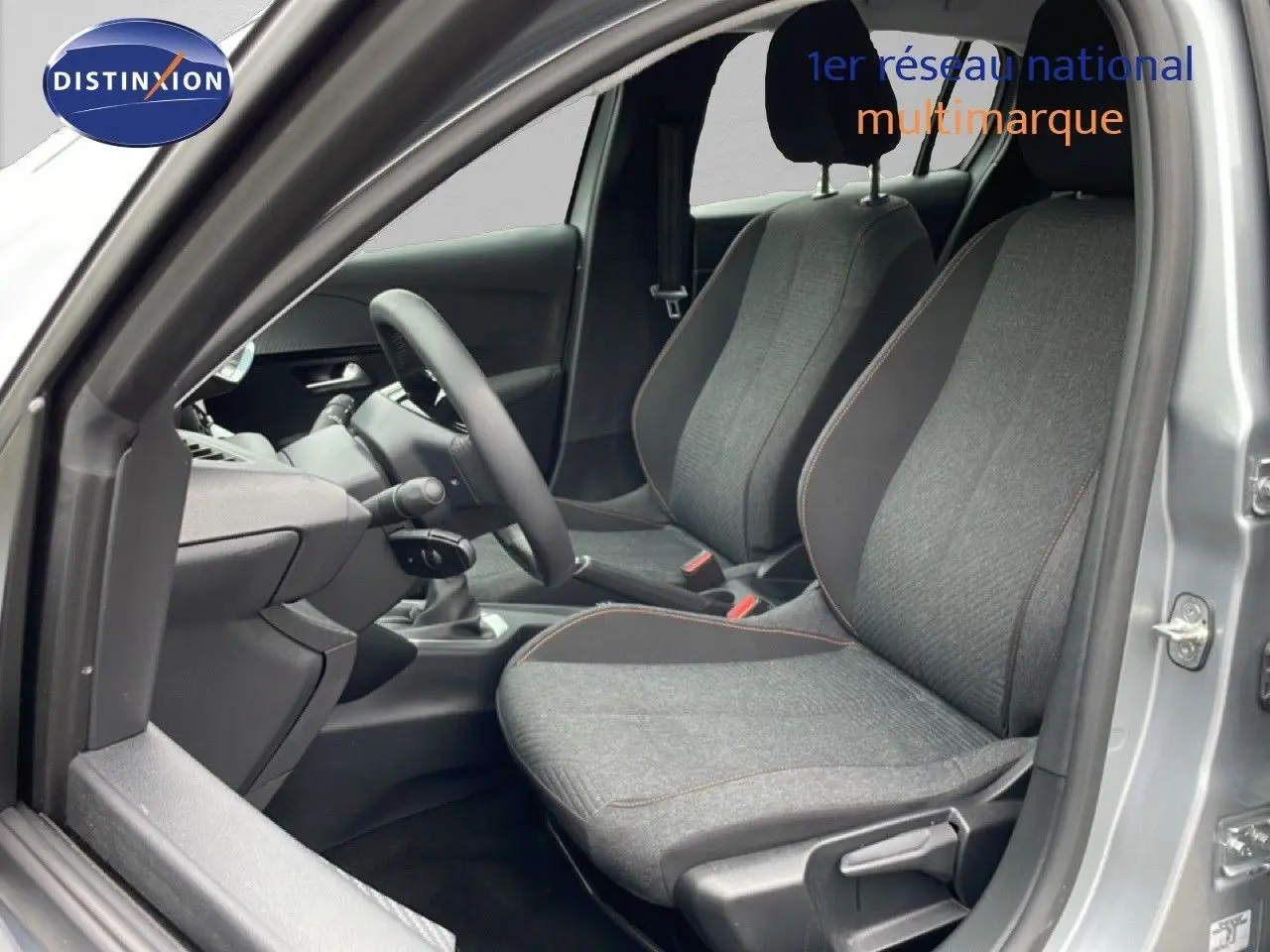 Vue intérieure côté conducteur de la Peugeot 208 gris Artense métal, sièges tissu gris avec surpiqûres orange.