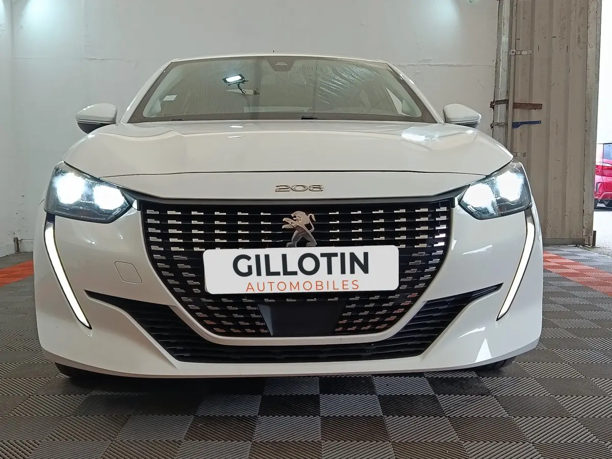 Vue avant d'une Peugeot 208 blanche de 2021 avec phares LED allumés et calandre distinctive noire.