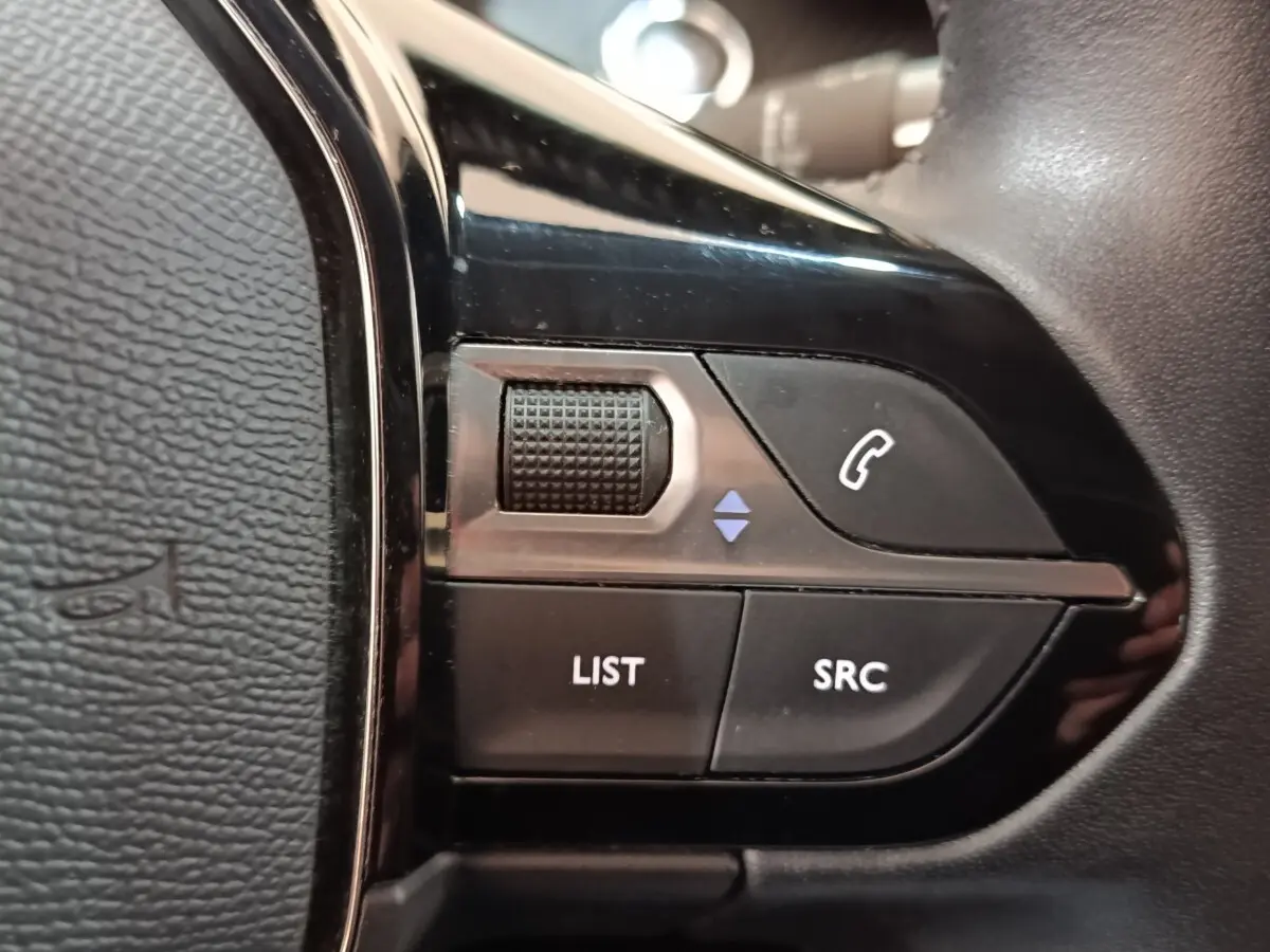 Gros plan sur les commandes noires du volant côté gauche d'une Peugeot 208 blanche, version PureTech 100 S&S Allure Business.