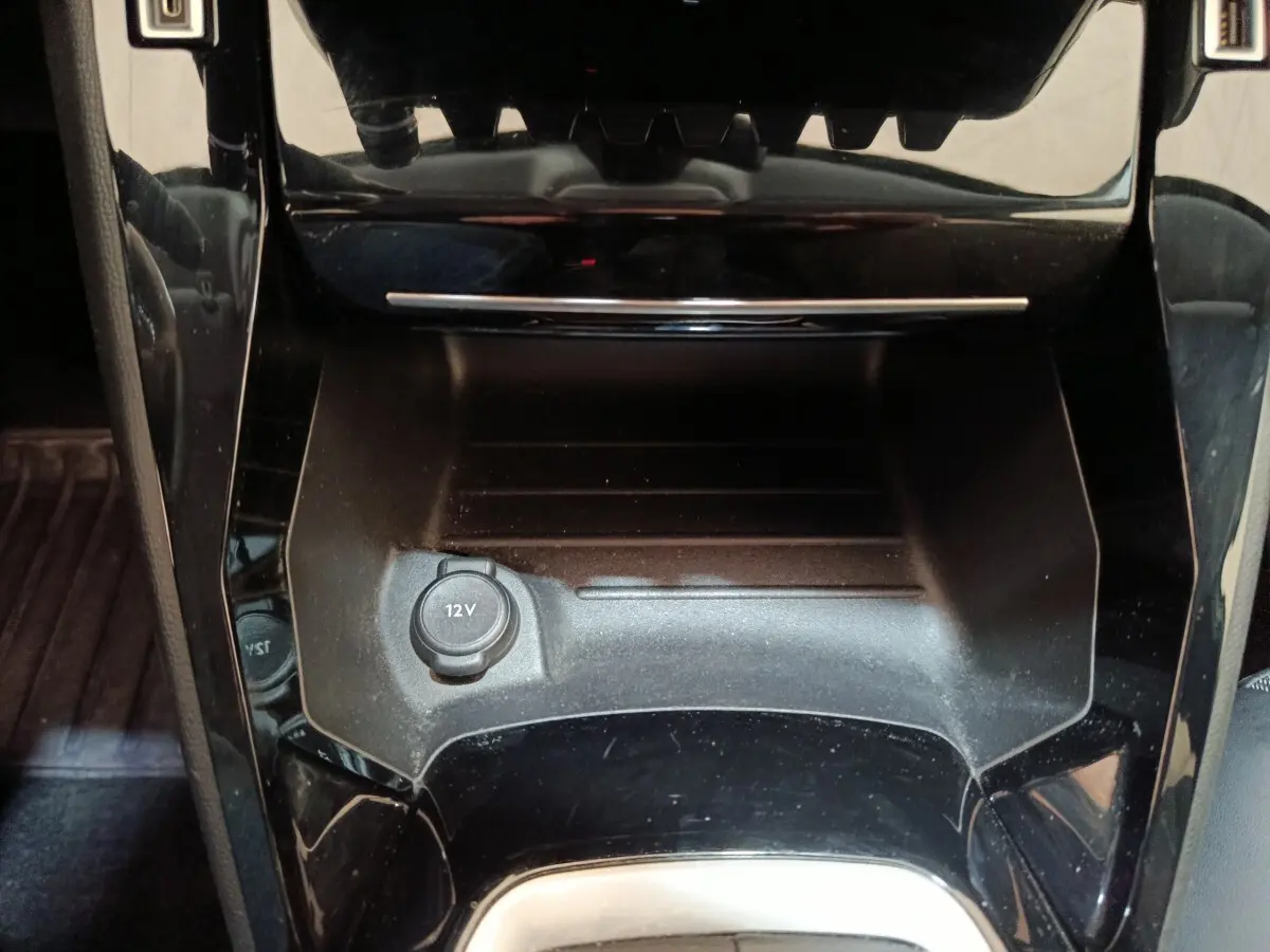 Gros plan sur la prise 12V et le rangement central noir brillant de la Peugeot 208 blanche, intérieur avant.