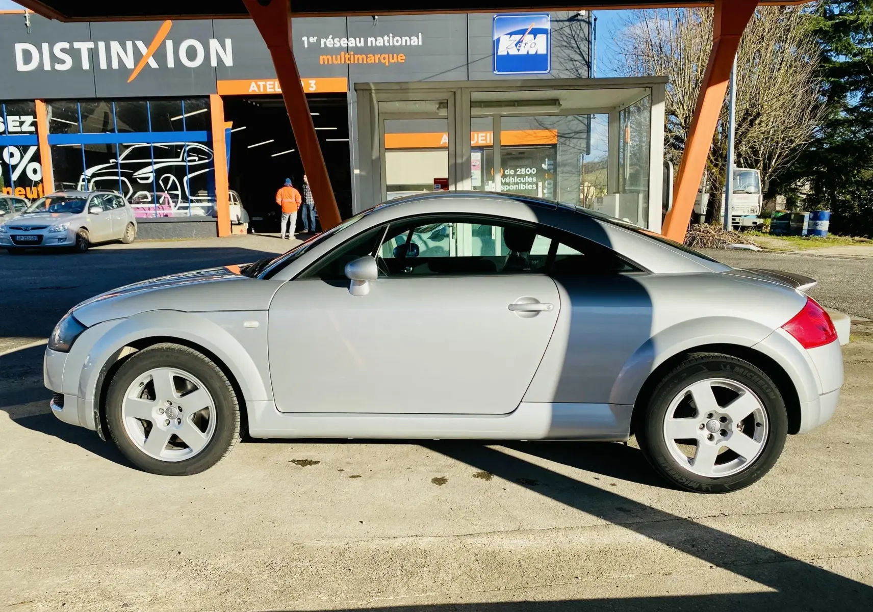 Profil côté gauche d'une Audi TT Coupé gris 2002 garée devant un garage avec ombre portée sur la carrosserie.