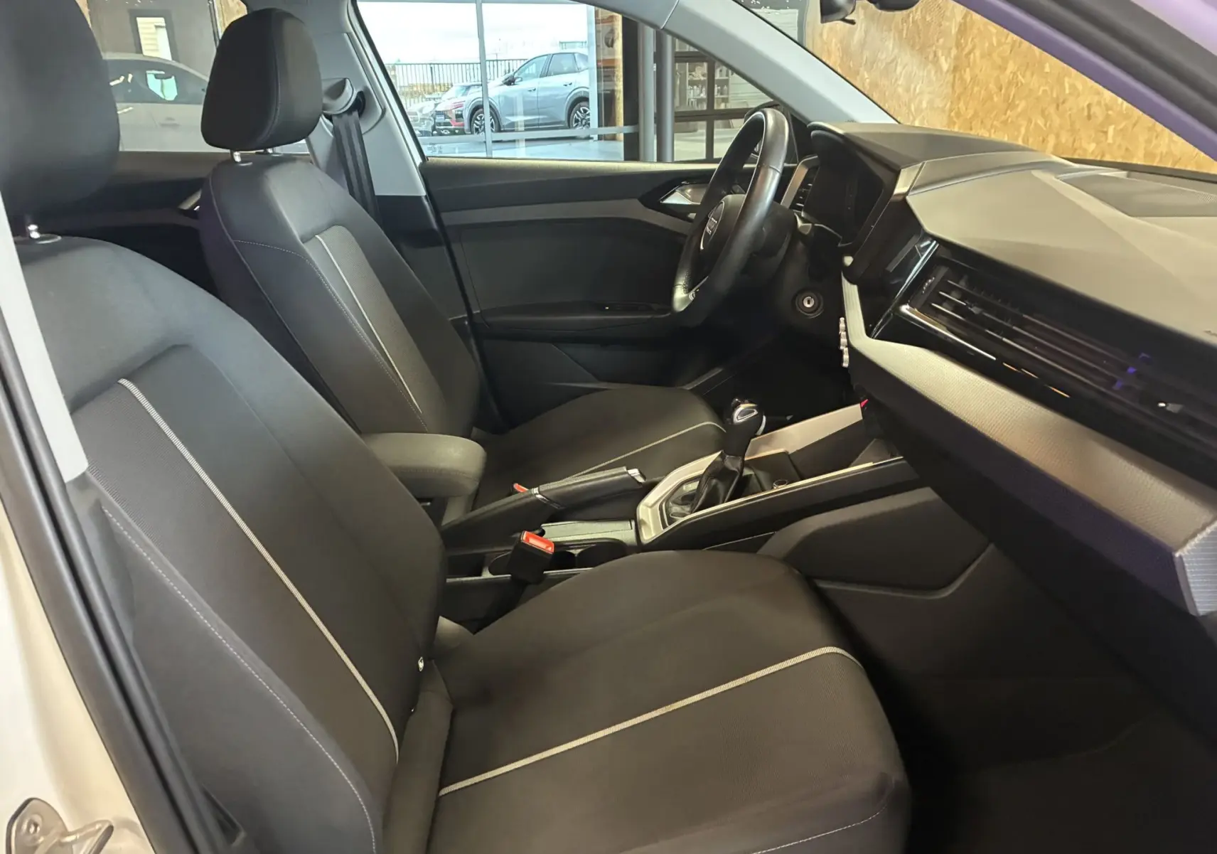 Vue intérieure côté gauche de l'Audi A1 blanche, montrant les sièges tissu noirs et le tableau de bord moderne.