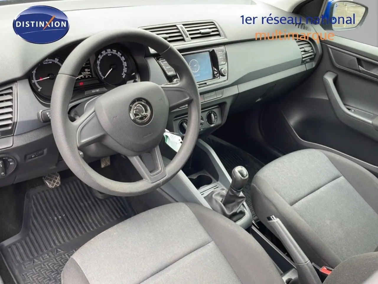 Vue intérieure côté conducteur de la Skoda Fabia 2019, tableau de bord noir et sièges tissu gris, volant et levier de vitesse visibles.