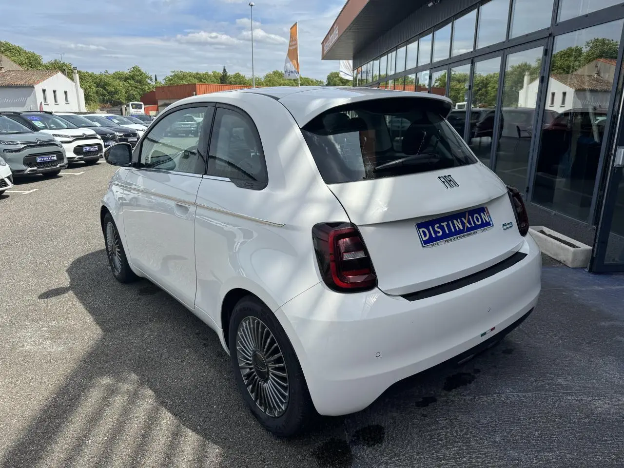 Vue 3/4 arrière droite d'une Fiat 500 électrique blanche avec jantes alliage et toit panoramique, stationnée devant un showroom.