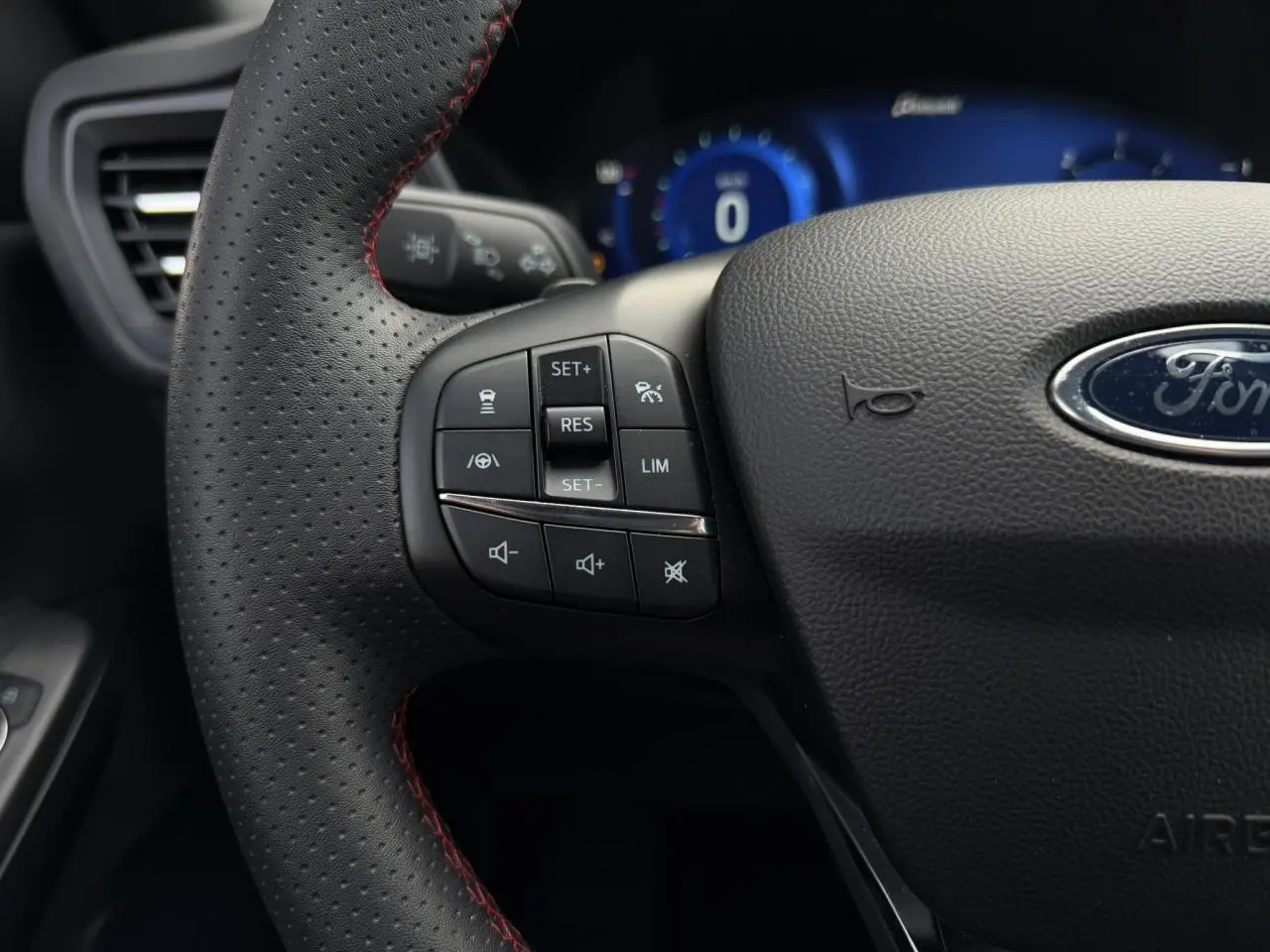 Gros plan sur le volant perforé noir avec surpiqûres rouges du Ford Kuga 2023, boutons de commande et tableau de bord numérique bleu en arrière-plan.