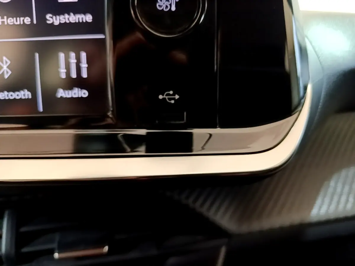 Gros plan sur la console centrale noire brillante de la Peugeot 208 blanche, avec port USB et écran tactile partiel visible.