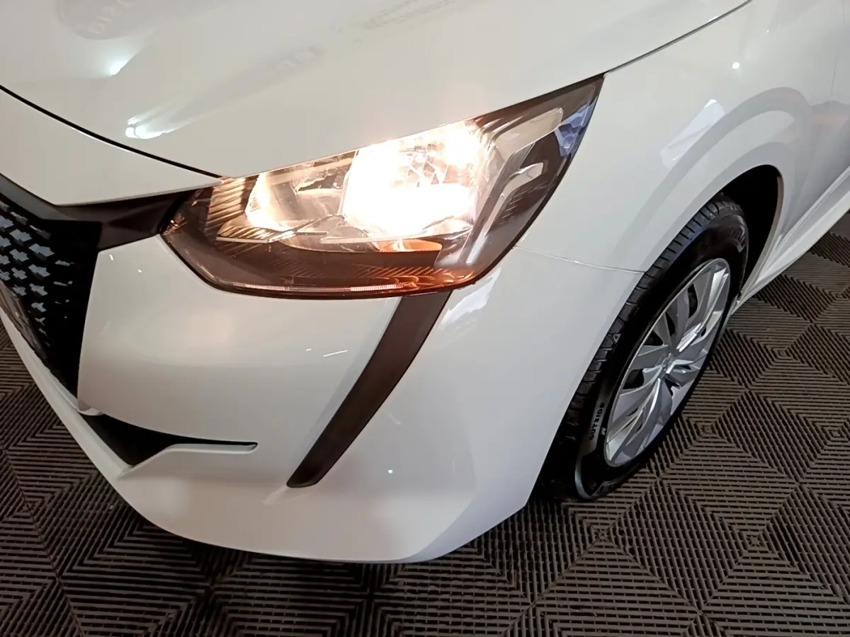 Gros plan sur l'avant droit blanc d'une Peugeot 208 2022 avec phare allumé et jante visible sur sol texturé.