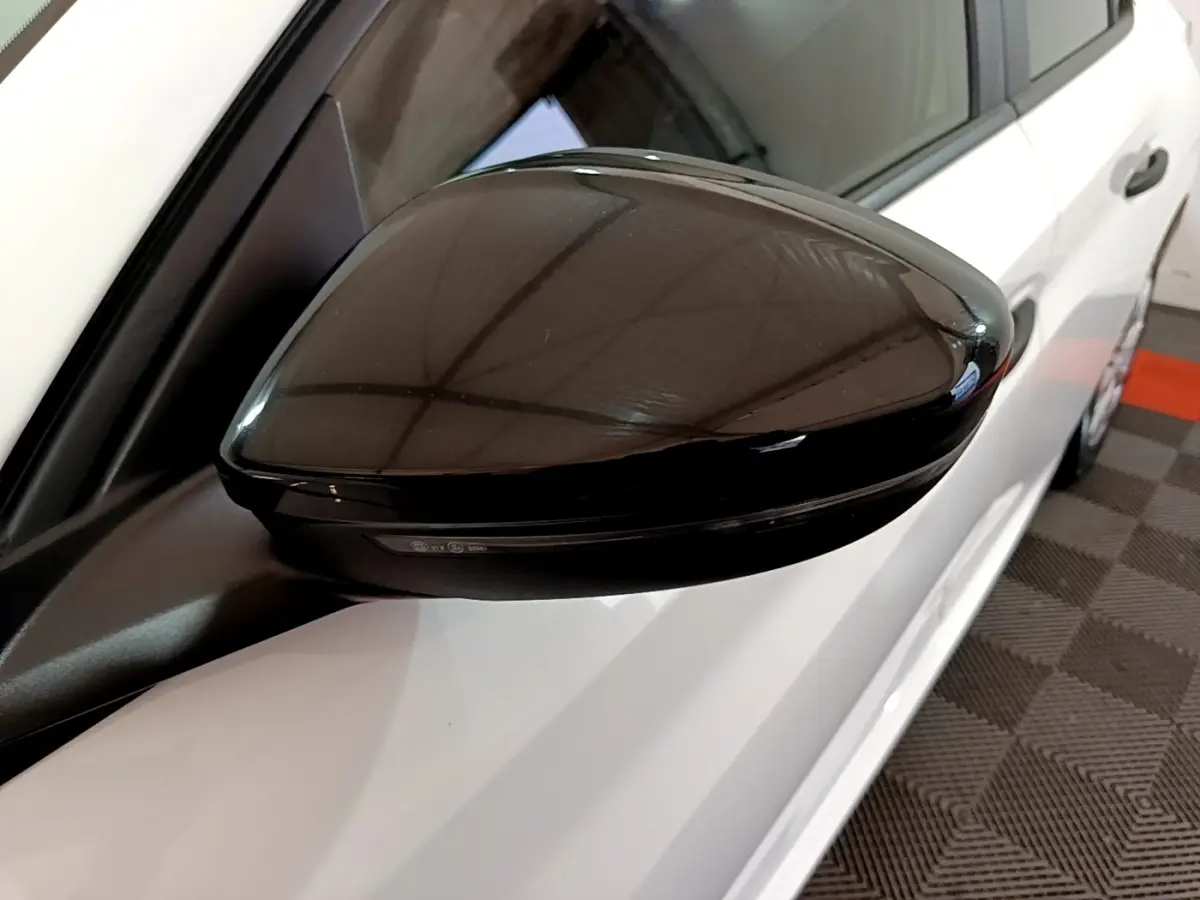 Gros plan sur le rétroviseur noir côté gauche d'une Peugeot 208 blanche vue de profil, stationnée en intérieur.