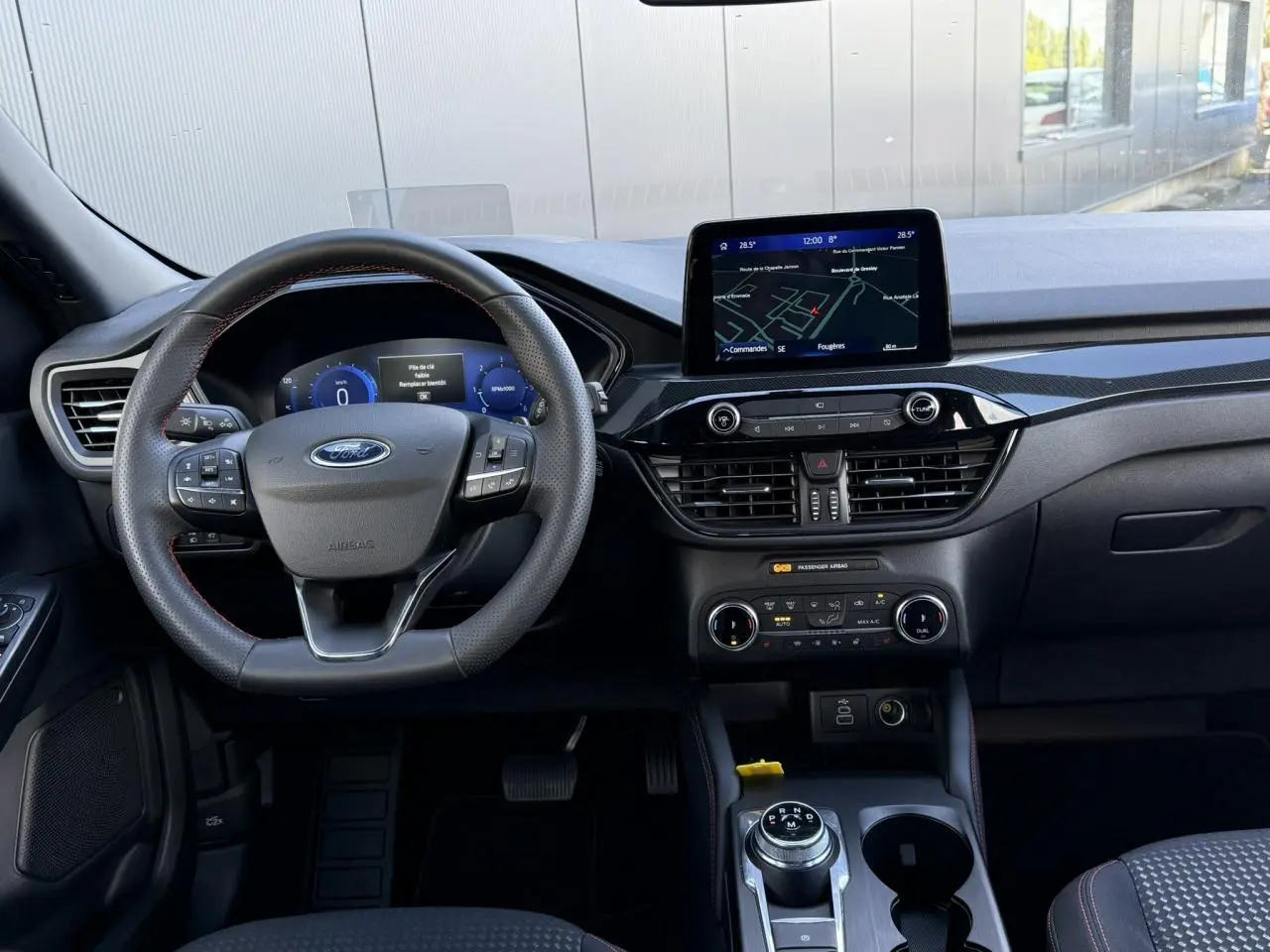 Vue intérieure avant du Ford Kuga 2024 ST-Line, tableau de bord noir avec écran tactile et volant à méplat avec surpiqûres rouges.