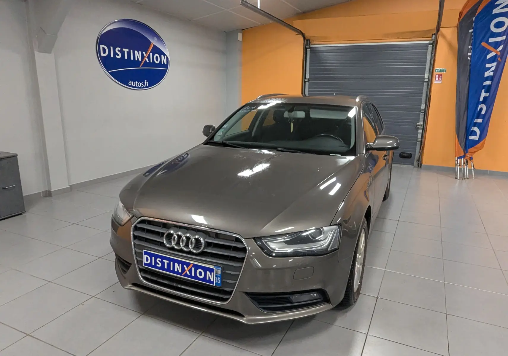 Audi A4 gris 3/4 avant droit en intérieur, avec plaque et signalétique Distinxion visibles sur le mur et au sol.