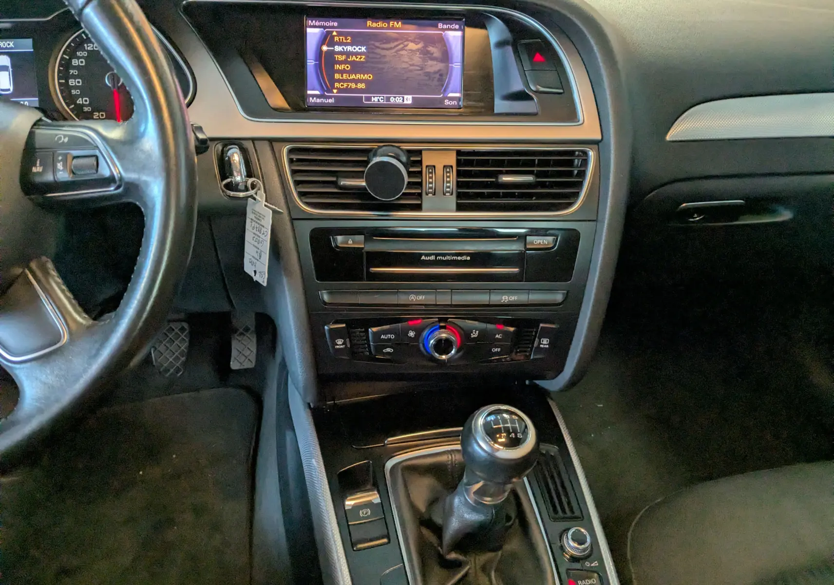 Vue intérieure centrée sur la console centrale et levier de vitesse manuelle d'une Audi A4 gris 2013 avec écran multimédia allumé.