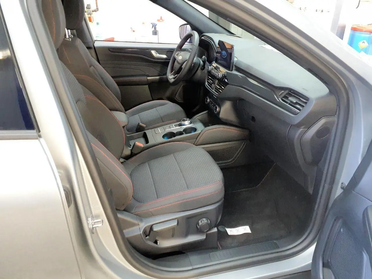 Vue intérieure côté conducteur du Ford Kuga gris Solar 2024, sièges noirs avec surpiqûres rouges et console centrale moderne.