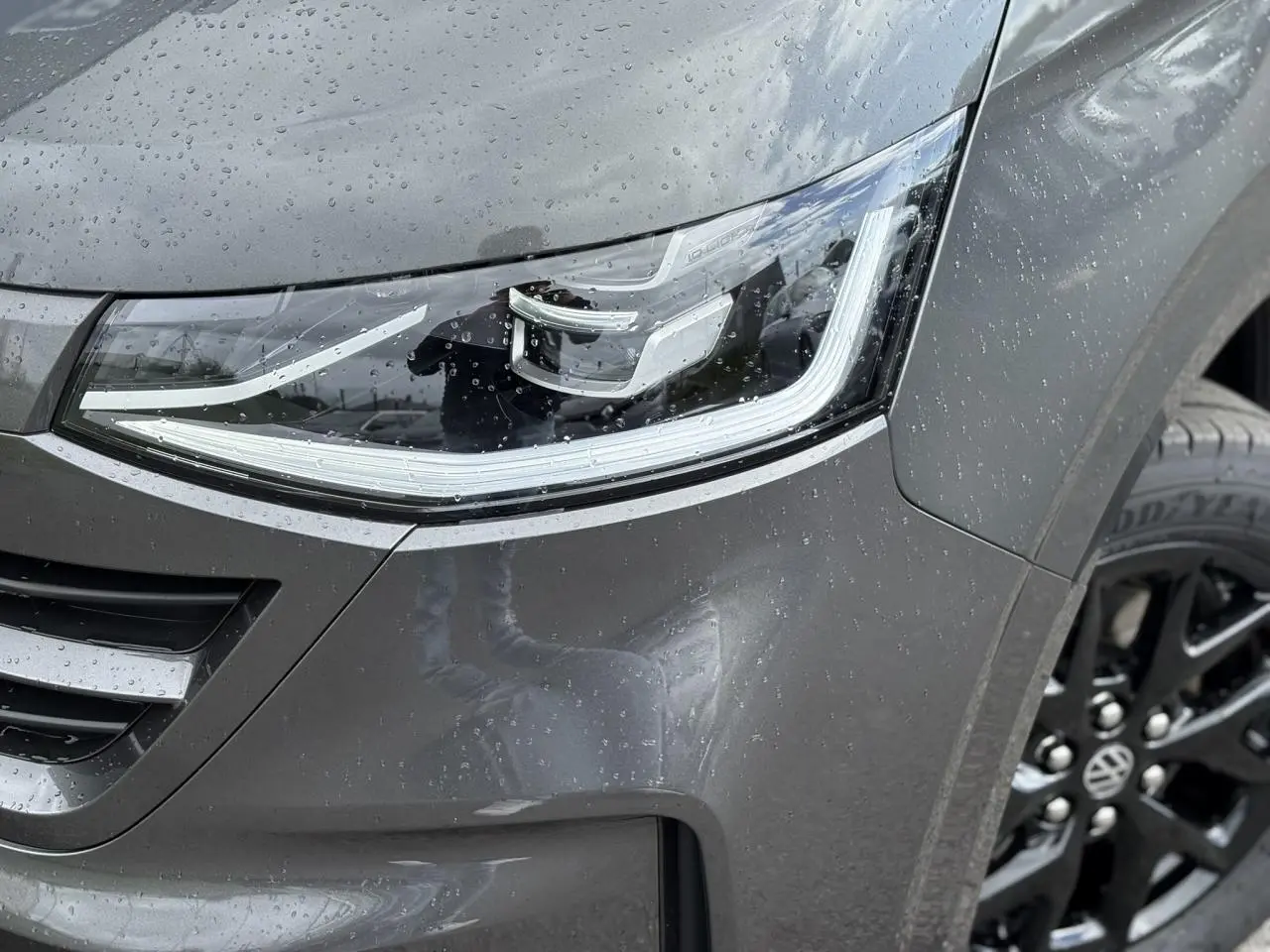 Gros plan sur le phare avant droit LED et la jante alliage noir brillant du Volkswagen Transporter T7 gris graphite mouillé.