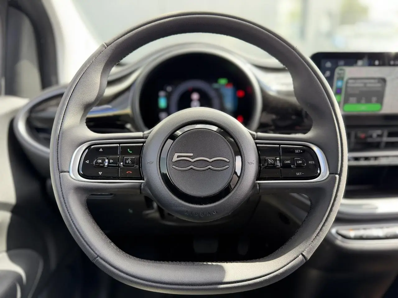 Vue rapprochée du volant noir Soft-Touch du Fiat 500 électrique 2024 avec commandes intégrées et tableau de bord numérique flou.